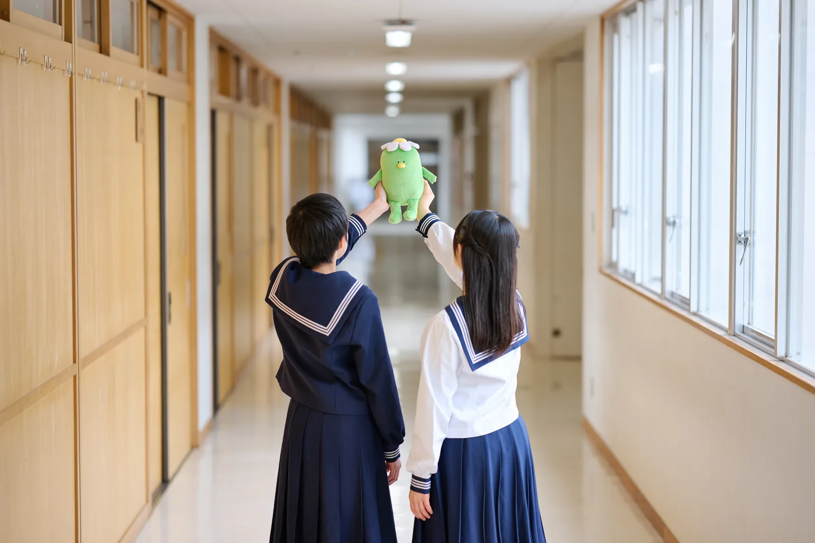 相模女子大学中学部・高等部の廊下で、生徒2人が後ろ姿で学園キャラクター「さがっぱ・ジョー」のぬいぐるみを高く掲げている