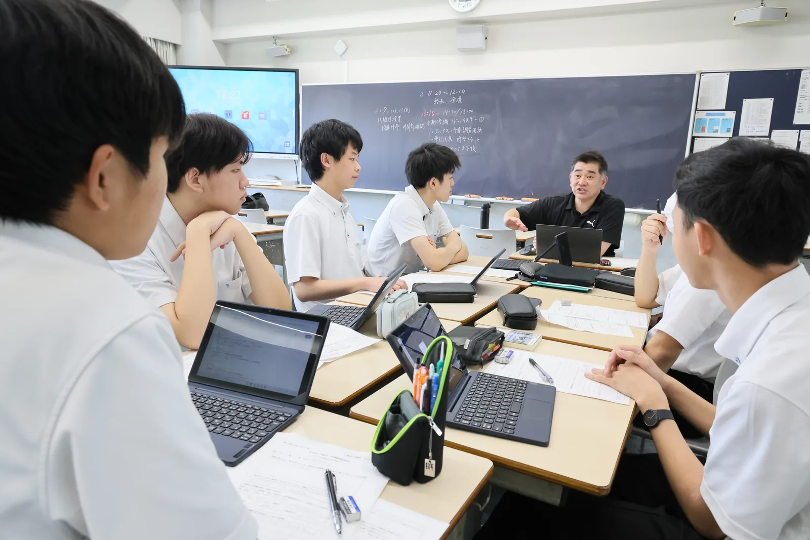 東京都市大学付属中学校・高等学校の教室での学習の様子。生徒がノートパソコンを開いて作業し、黒板の前でも話し合いが行われている